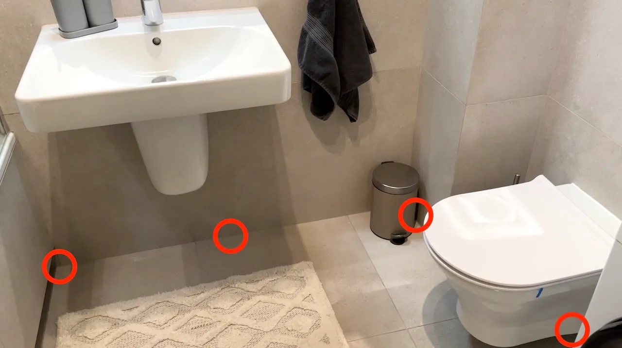 Łazienka z czterema czerwonymi kółkami: przy ścianie pod umywalką, na złączu płytek, obok kosza przy toalecie i u podstawy muszli