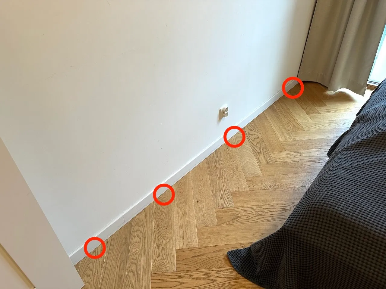 Listwa przypodłogowa w sypialni z czterema czerwonymi kółkami oznaczającymi miejsca aplikacji żelu wzdłuż ściany, podłoga w jodełkę