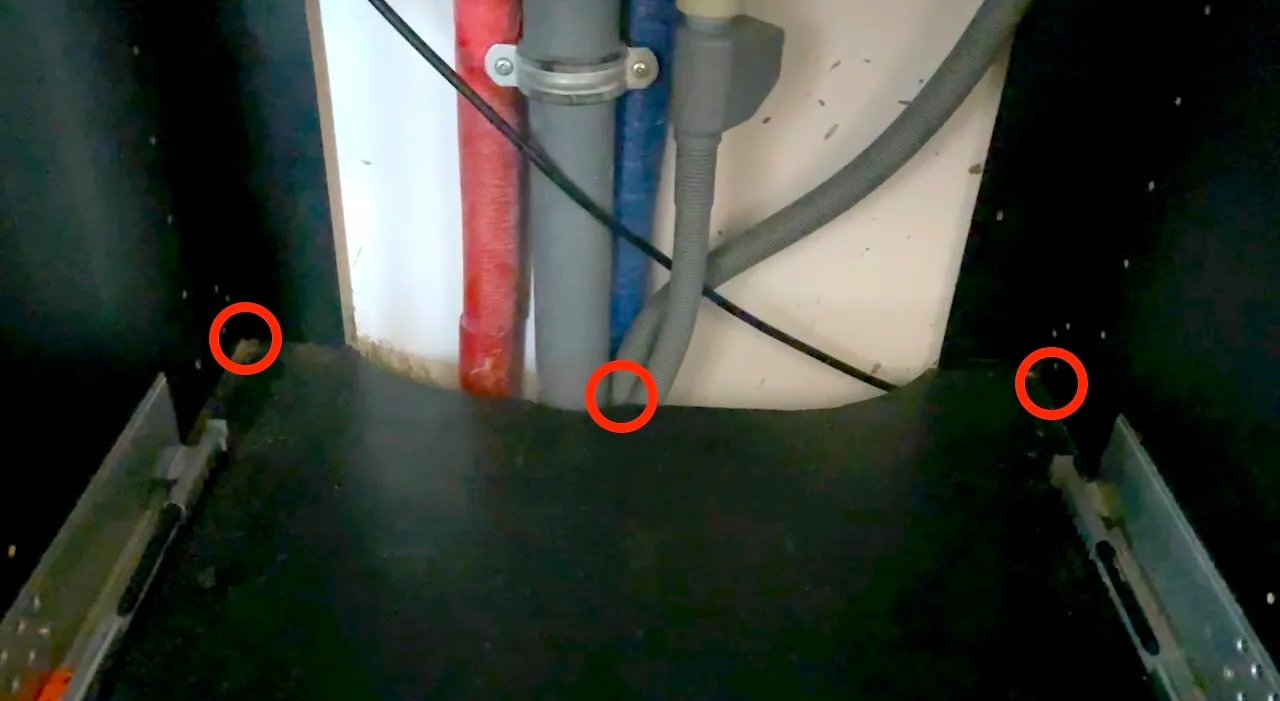 Wnętrze szafki pod zlewem z wyjętą szufladą, widoczne rury i wąż odpływowy, trzy czerwone kółka w narożnikach i przy przejściu węża